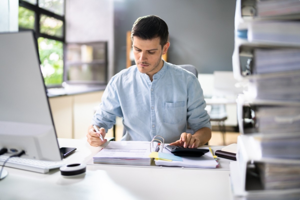 Man in light blue shirt working in bright office holding pen and using calculator 