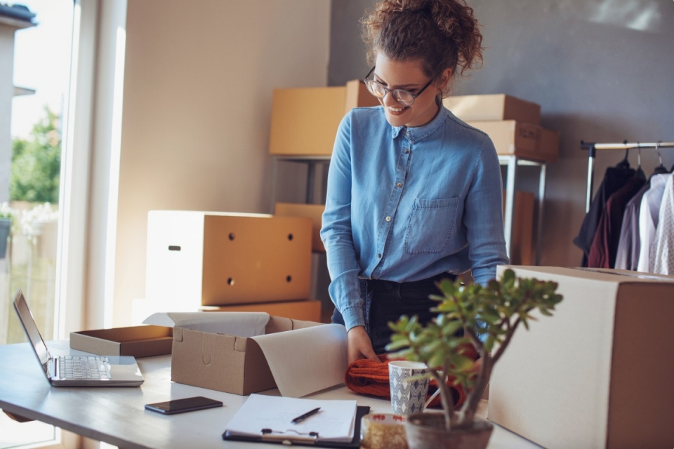Woman in blue shirt packing goods into box in a small storeroom with clothes on a rail in the background