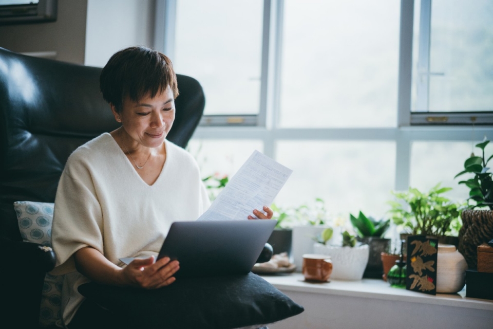 Middle-aged casually smart dressed business woman relaxed looking at laptop and holding paperwork
