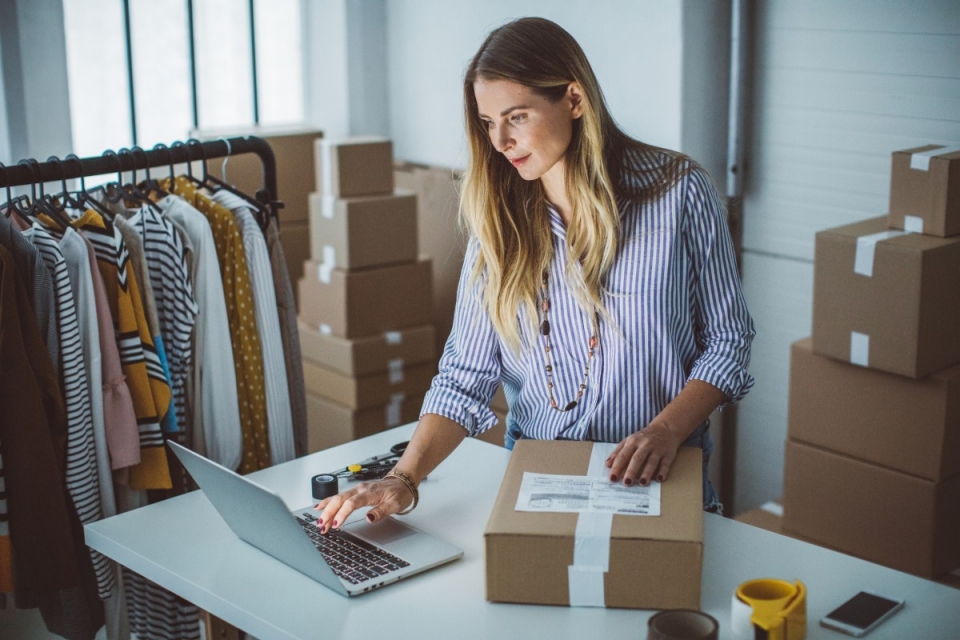 Female fashion retailer in stockroom labelling parcel and using laptop