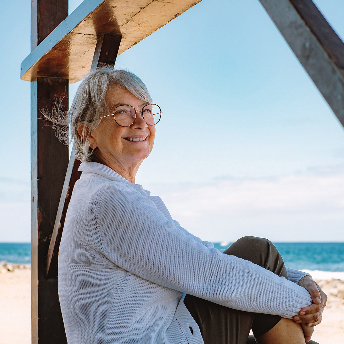 A grey haired woman smiling as she sits on the beach under a pier